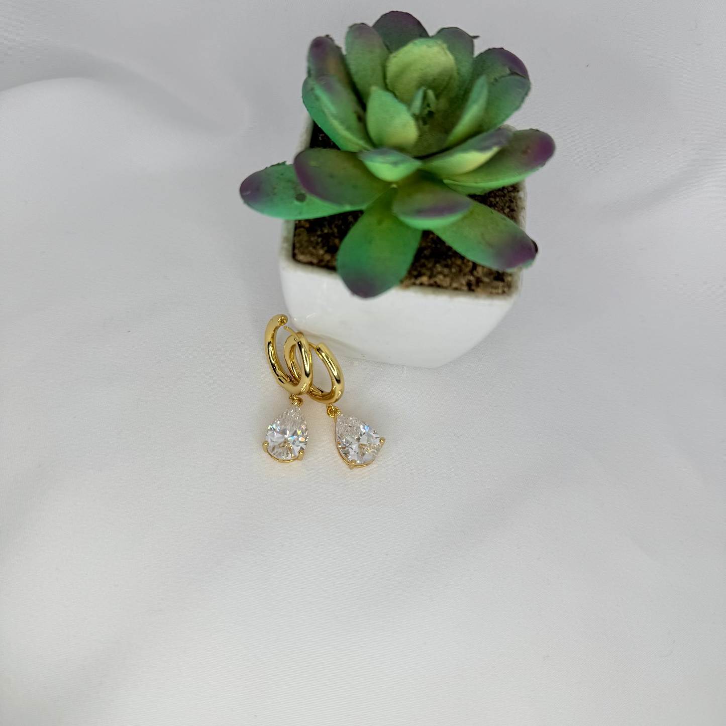 Gold earrings with clear stones on a white surface with a small potted succulent.