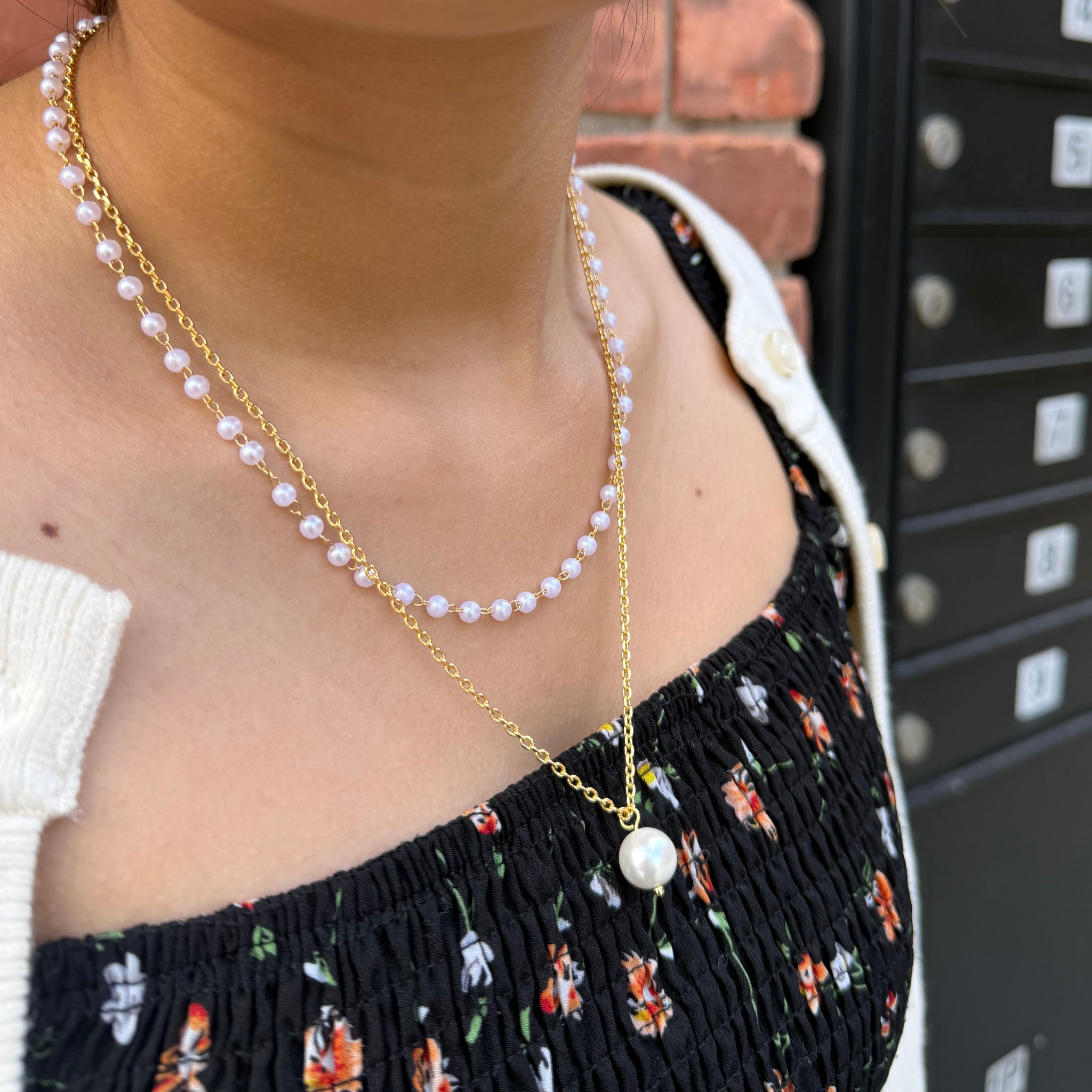 Person wearing a gold chain with pearls around their neck, sitting on a bench.