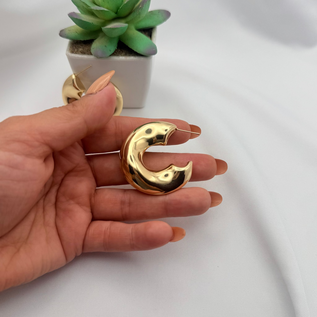Gold crescent moon earring held by a hand with a small potted plant in the background.