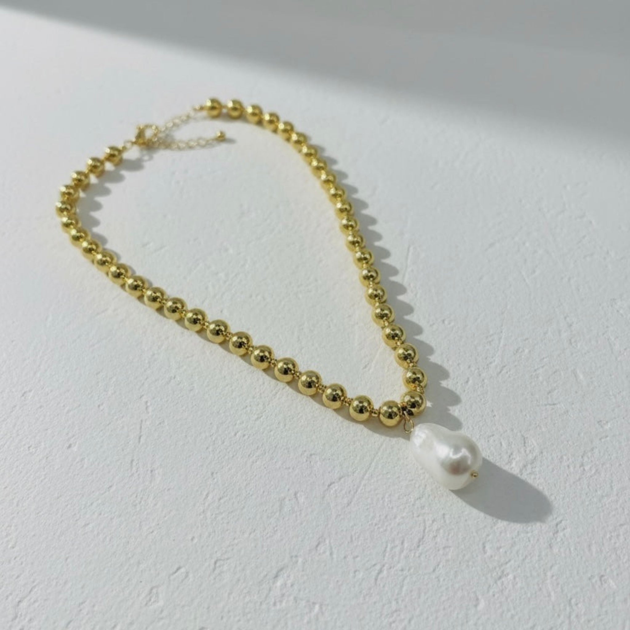 Gold beaded necklace with a single pearl on a light gray background