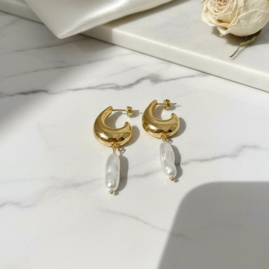 Gold hoop earrings with clear beads on a marble surface with a white flower and drawstring bag.