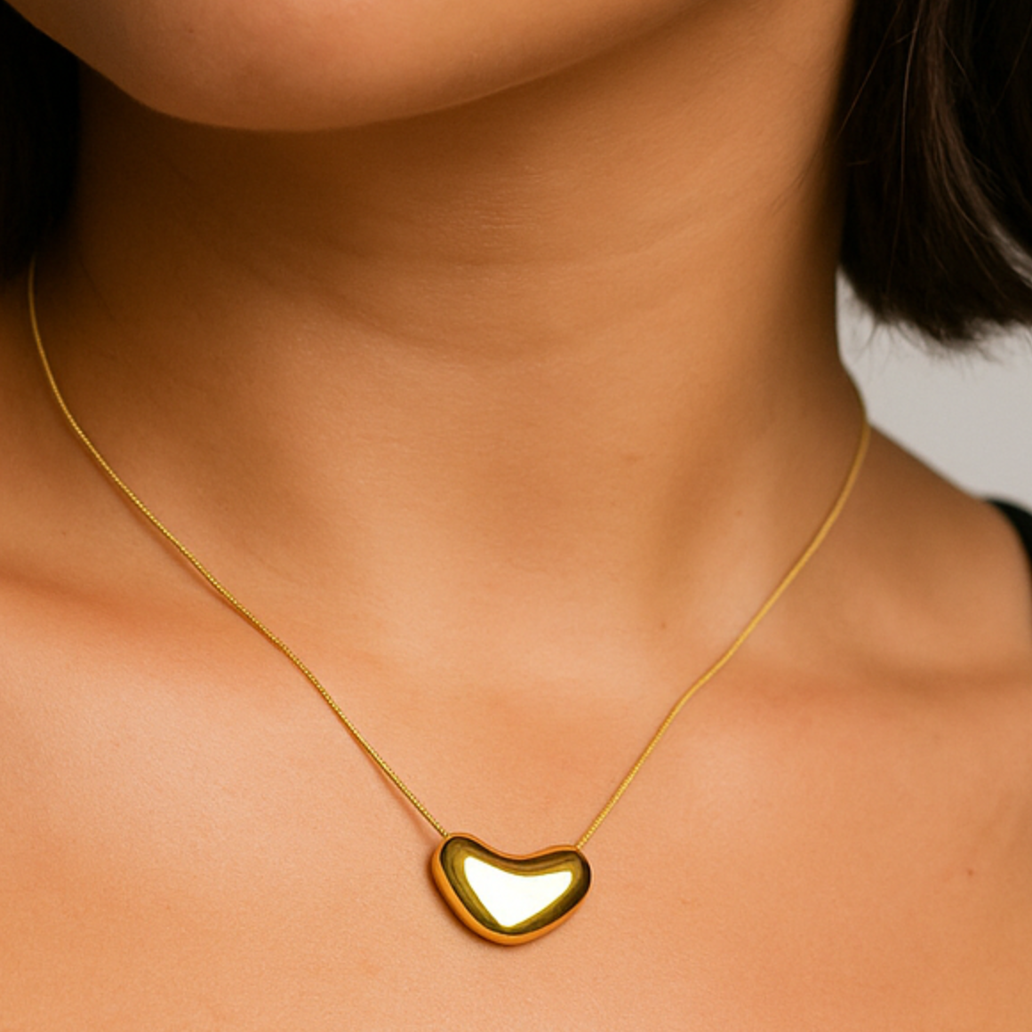 Close-up of a person wearing a gold heart-shaped necklace against a neutral background