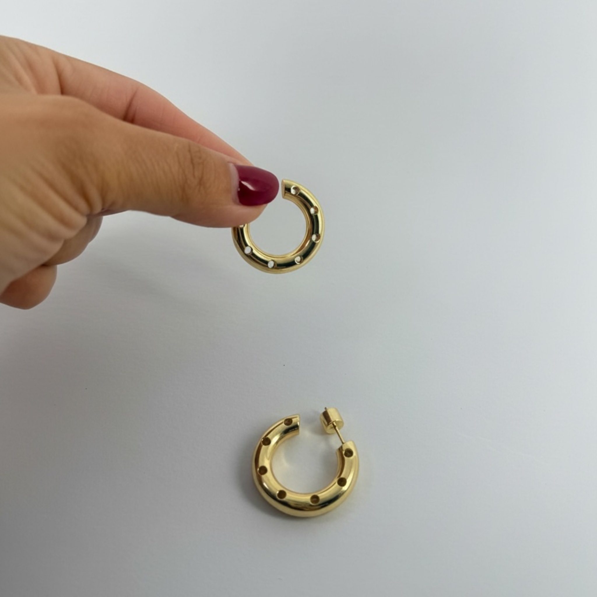 Gold hoop earrings held by a hand with red nail polish on a light gray background