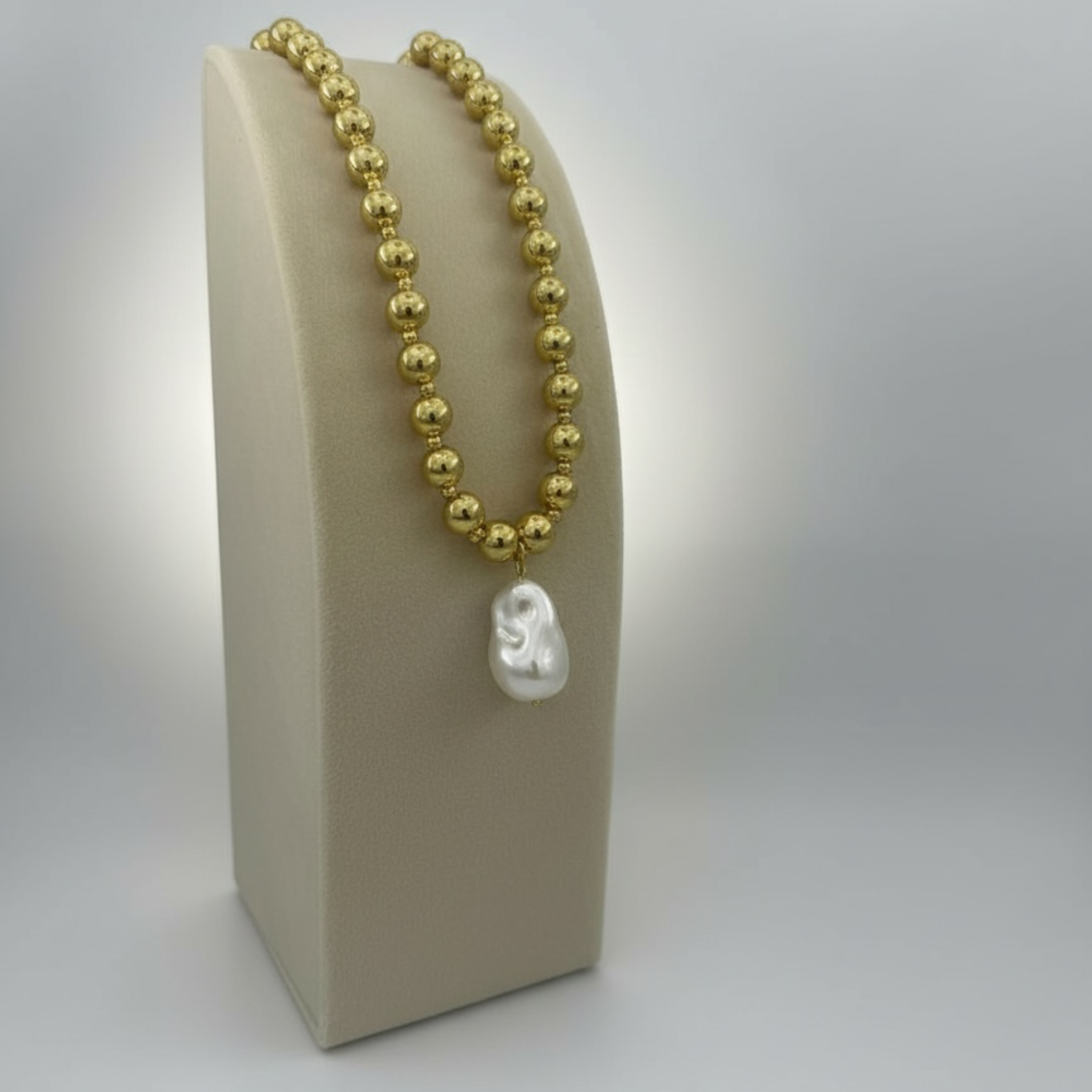 Gold beaded necklace with a white pendant on a beige stand against a light gray background